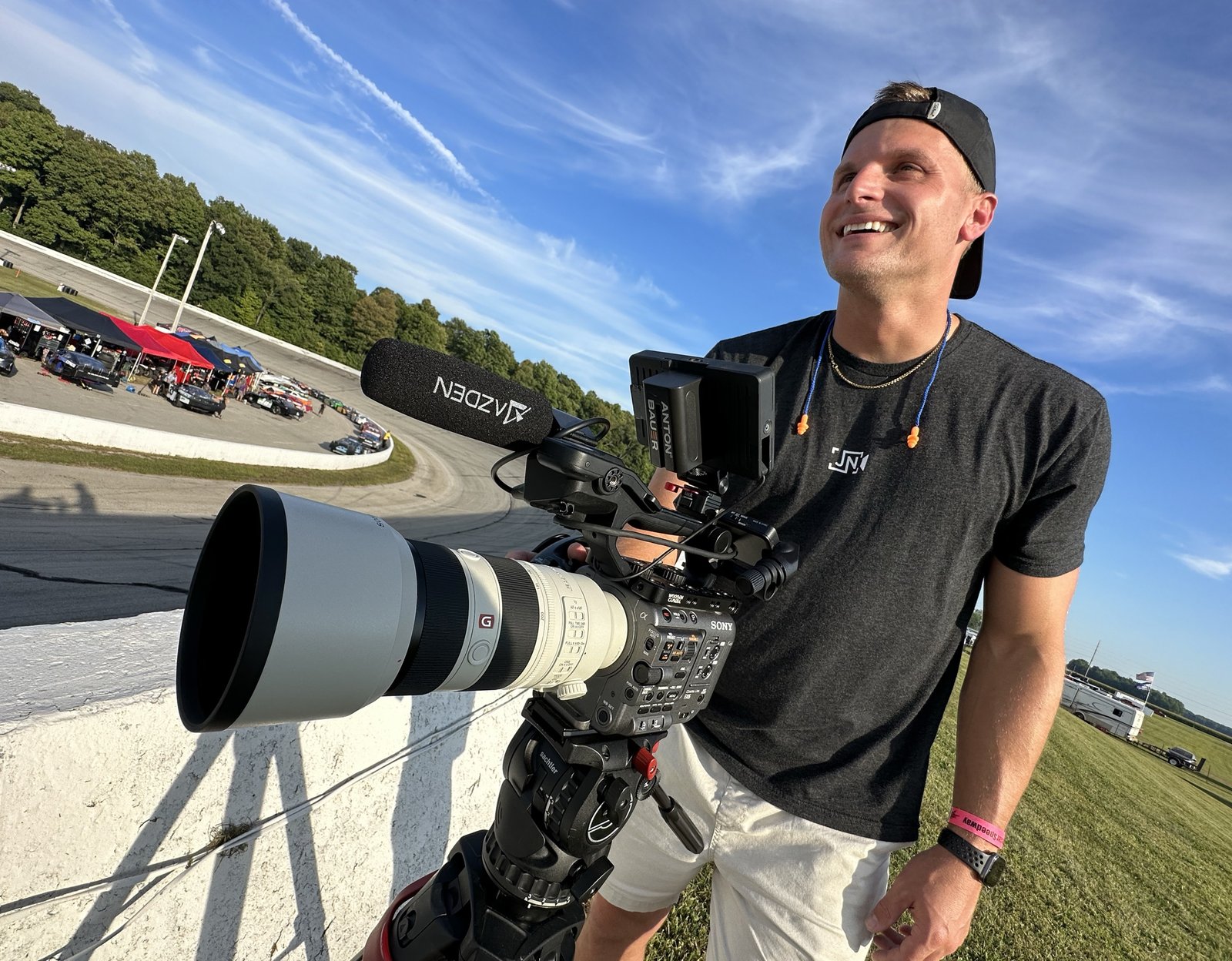 Josh Nagel smiling while operating a Sony camera rig trackside at a speedway, with race cars visible in the background.